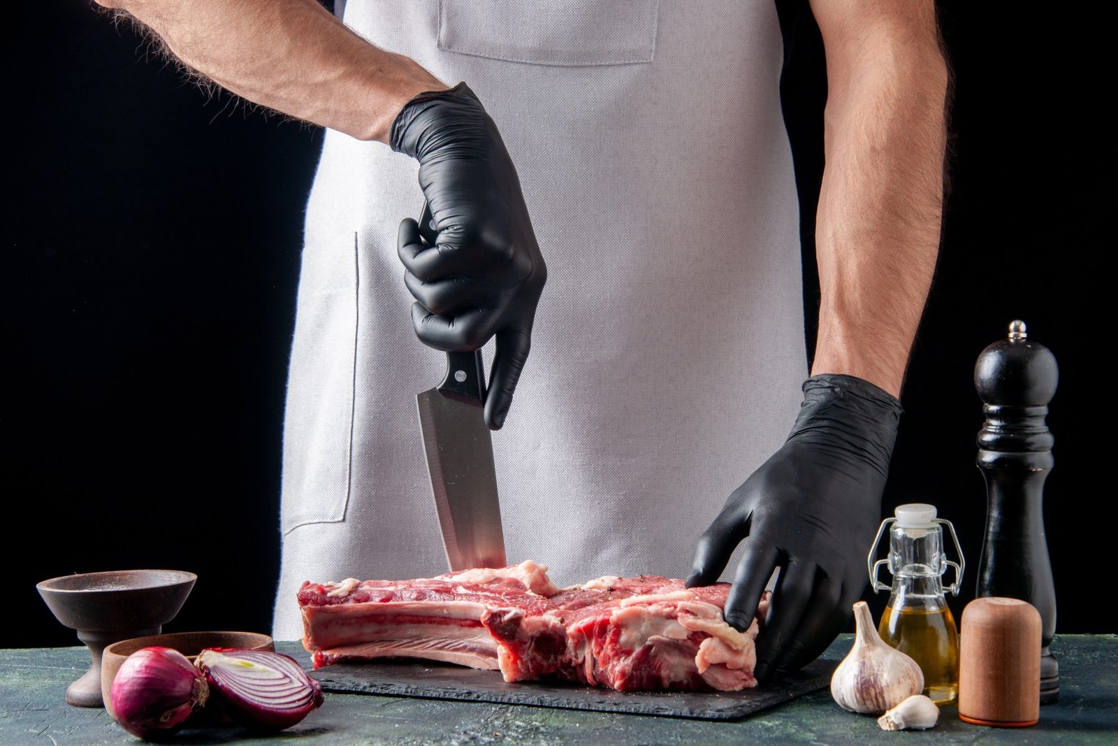 A professional butcher wearing black gloves uses a large knife to portion a thick piece of raw, bone-in beef.