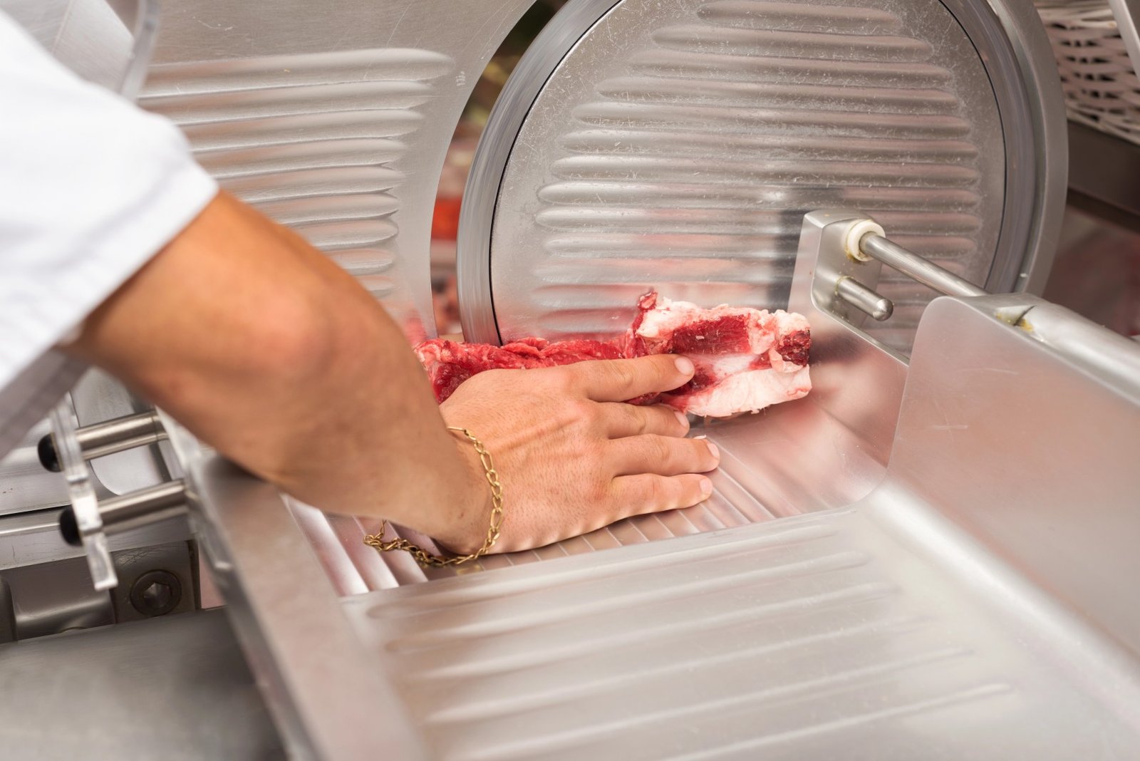 A butcher's hand guides a piece of raw beef through a commercial stainless steel meat slicer for uniform cuts.