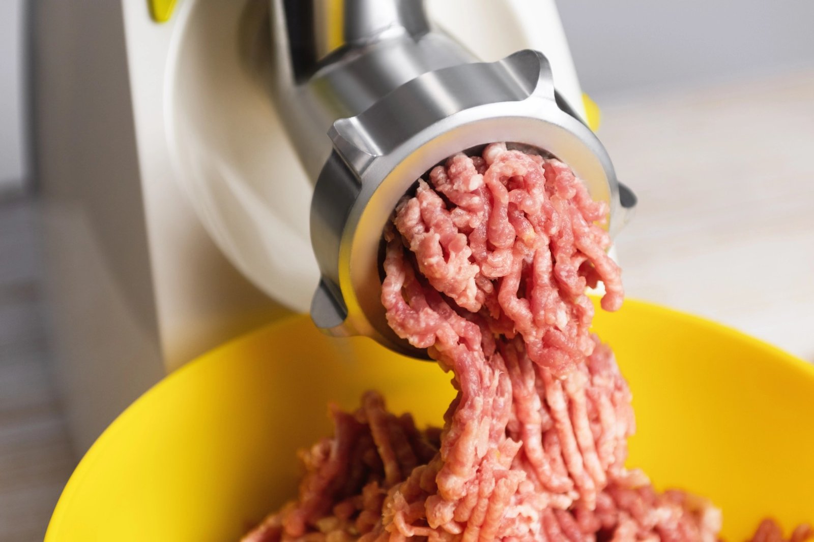 Freshly ground raw beef extrudes from a stainless steel meat grinder into a bright yellow plastic bowl.