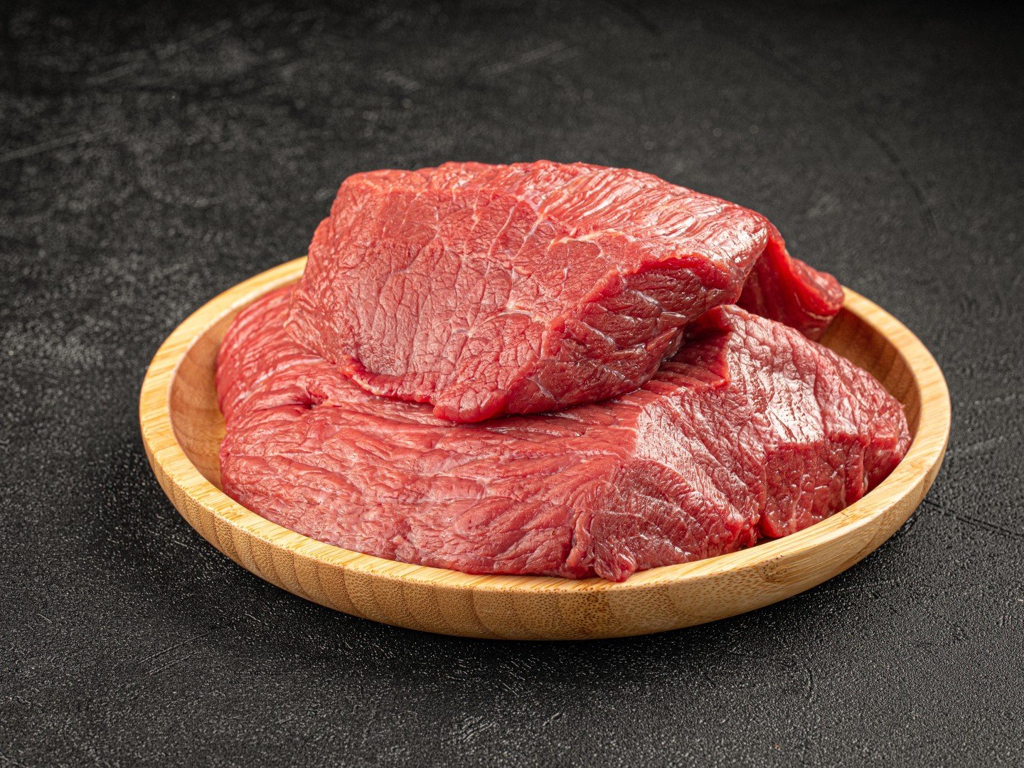 Several large, thick cuts of fresh raw beef stacked on a light wooden plate for kitchen prep.