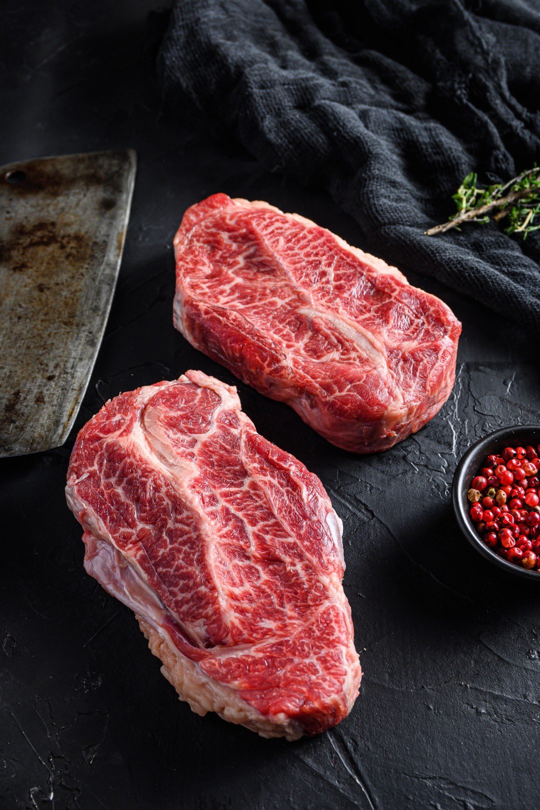 Two highly marbled raw top blade beef steaks next to a butcher cleaver on a dark slate surface.