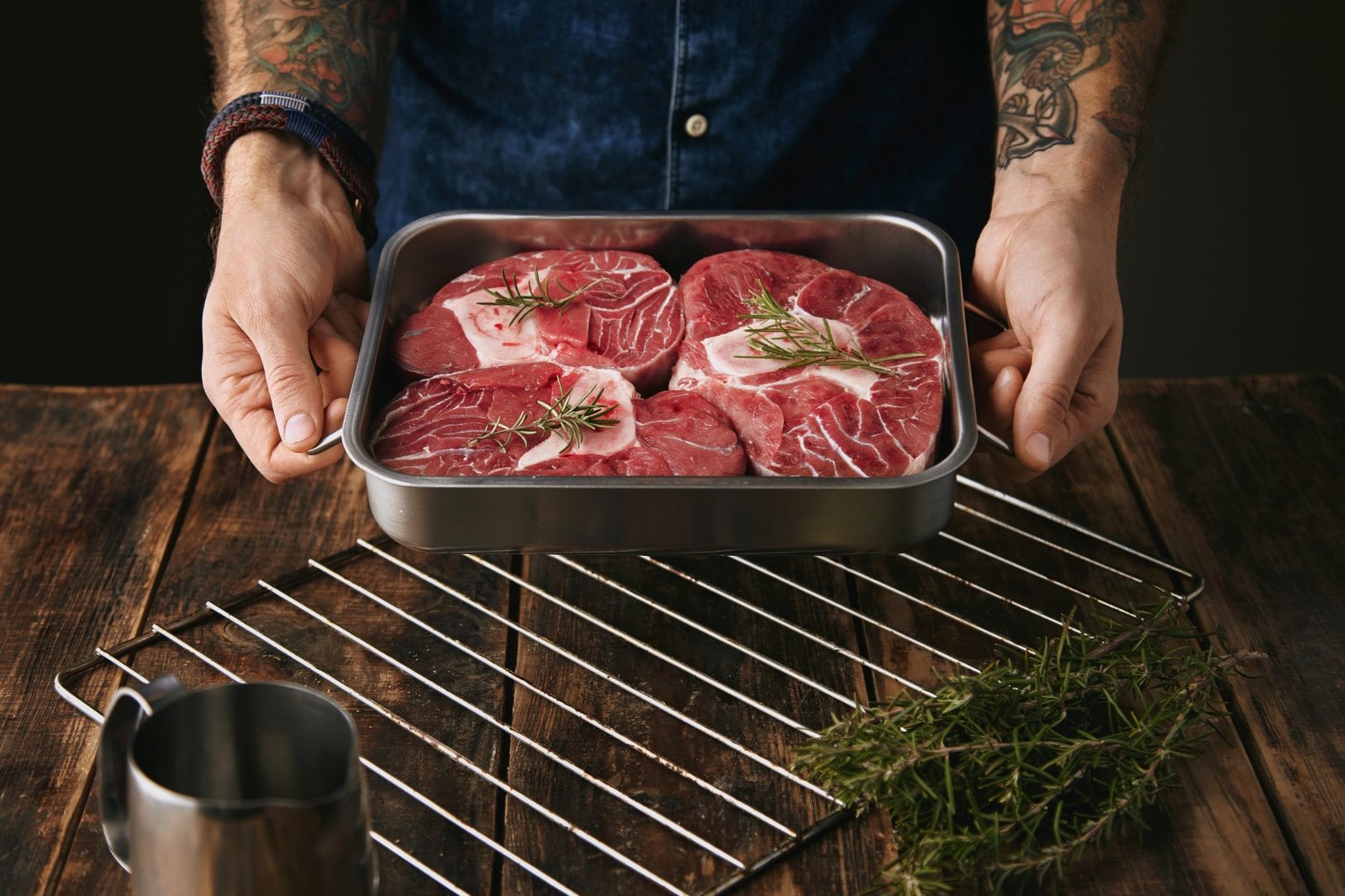 A person holding a stainless steel tray with three thick bone-in raw beef shanks garnished with rosemary.